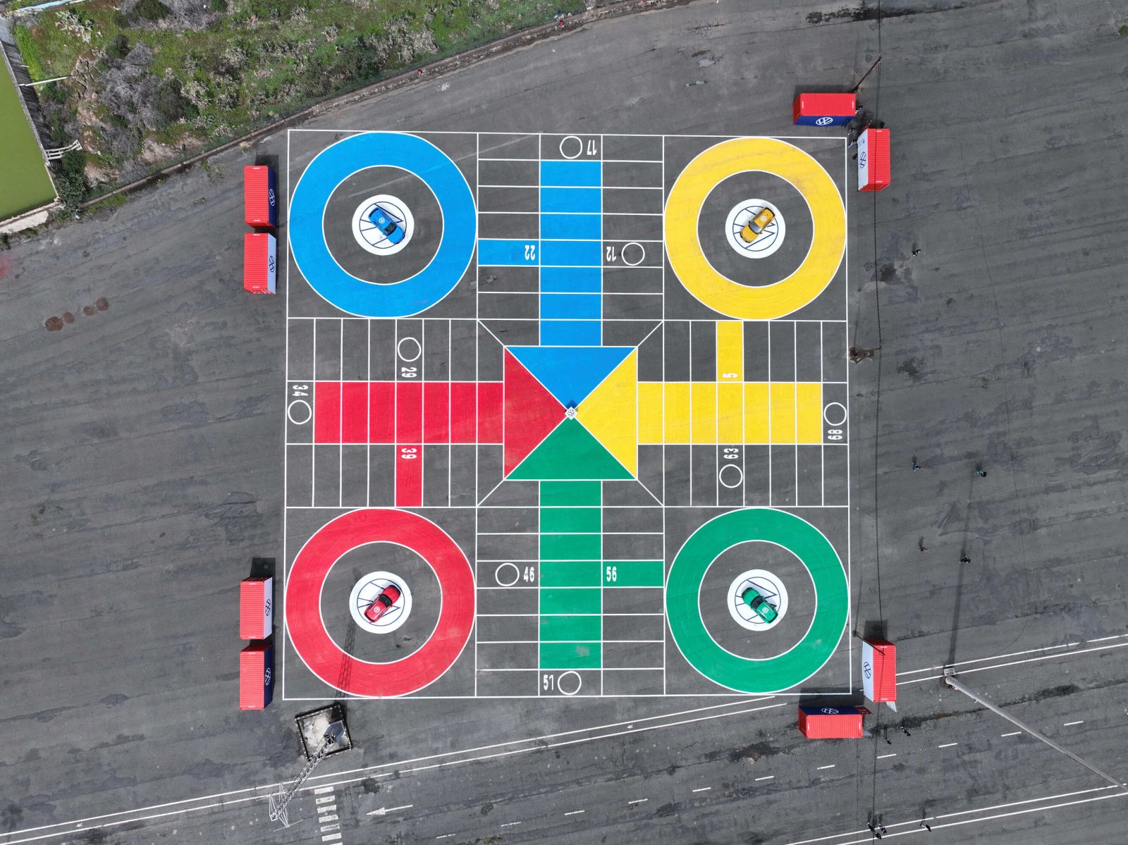 Vista aérea de un tablero gigante de Parchís pintado sobre una explanada de asfalto, con cuatro coches Volkswagen T-Roc de colores azul, amarillo, rojo y verde estacionados en sus respectivas casillas circulares de salida.